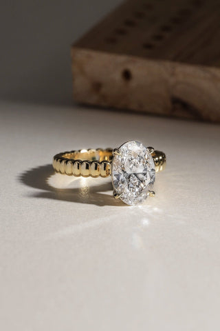 A yellow gold ring with a large oval-cut clear gemstone, resting on a light beige surface with a wooden block in the background.